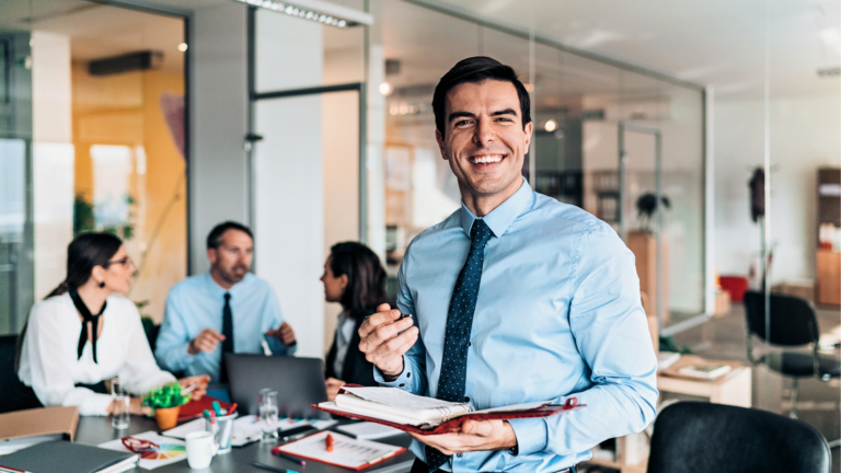 A man in a Suit Smiling while in the Background 3 other Sales Workers having a conversation