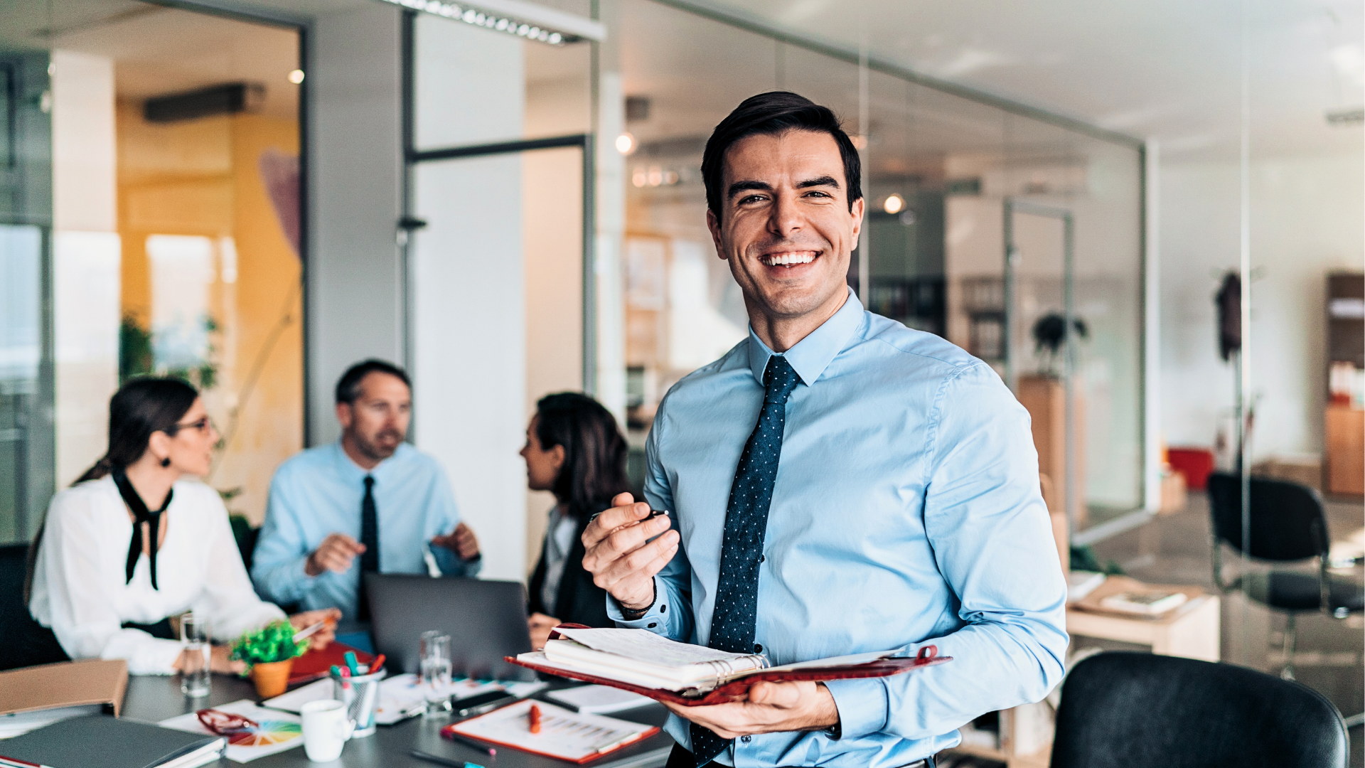 A man in a Suit Smiling while in the Background 3 other Sales Workers having a conversation