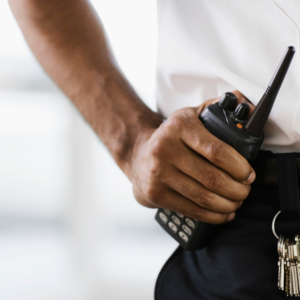 Nahaufnahme eines Sicherheitsdienstmitarbeiters in weißem Hemd, der ein Funkgerät in der Hand hält. Ein Schlüsselbund hängt an seinem Gürtel.