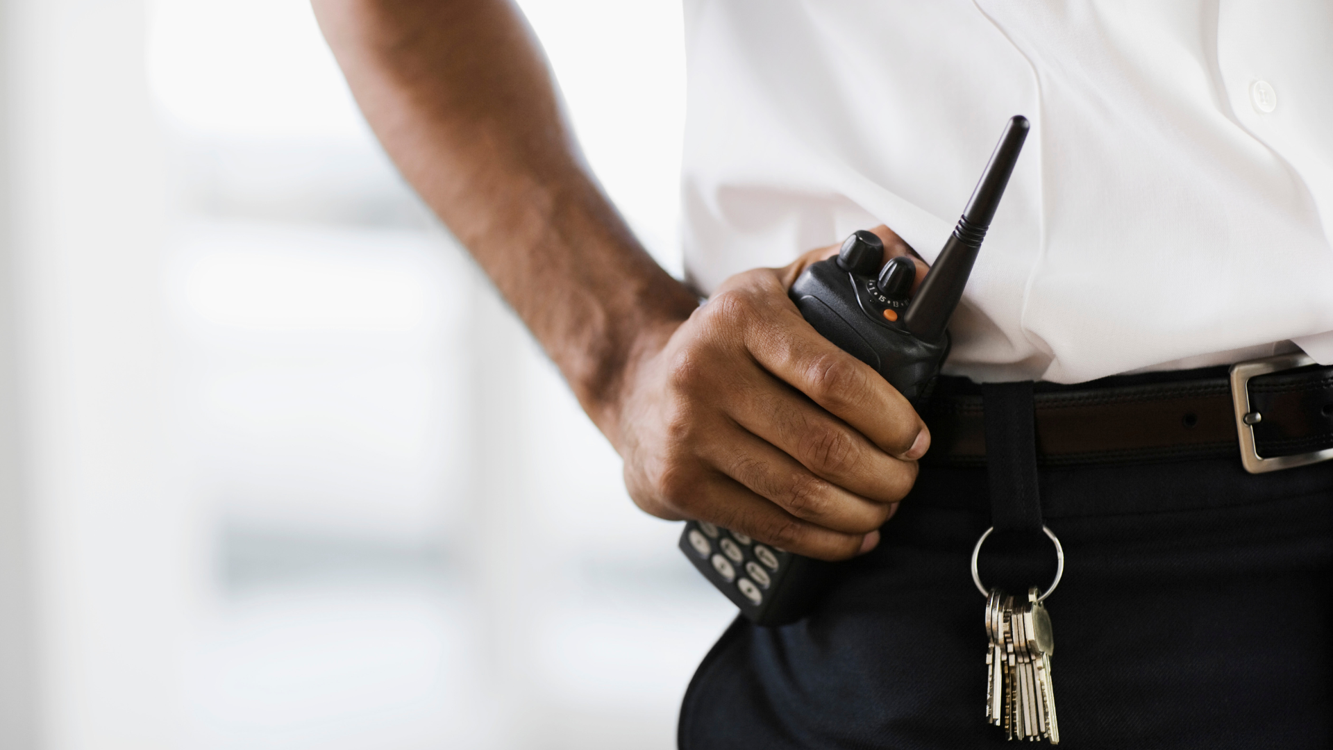 Nahaufnahme eines Sicherheitsdienstmitarbeiters in weißem Hemd, der ein Funkgerät in der Hand hält. Ein Schlüsselbund hängt an seinem Gürtel.