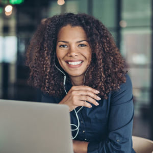 Frau mit lockigem Haar und Kopfhörern, die lächelnd an einem Laptop arbeitet, in einem modernen Büro.