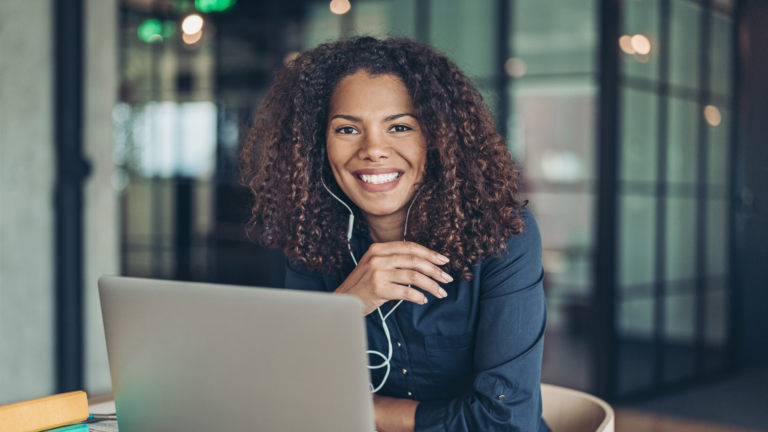 Frau mit lockigem Haar und Kopfhörern, die lächelnd an einem Laptop arbeitet, in einem modernen Büro.